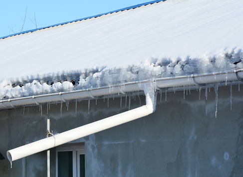Frozen gutters damage. House  roof covered snow, icicles and frozen roof gutter with downspout pipe.