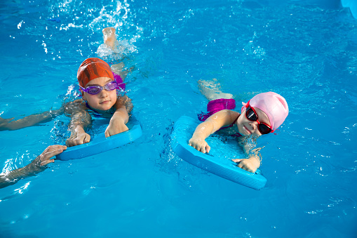 Two little girls learning how to swim in swimming pool and having fun using flutter boards