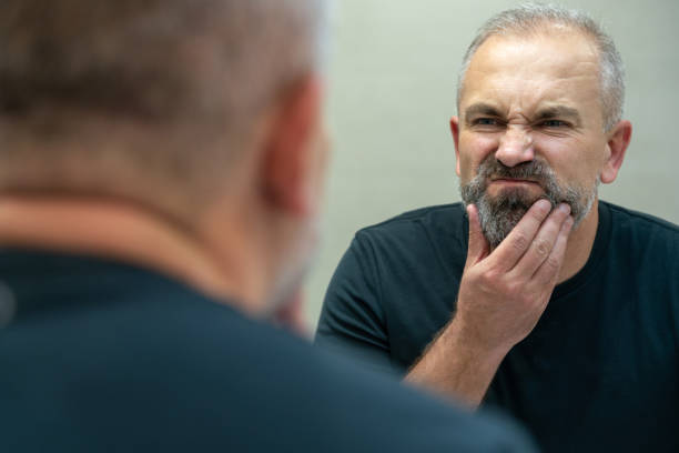istock Middle-aged handsome man looking in mirror in bathroom touching his beard thinking of cutting it off 1987249201