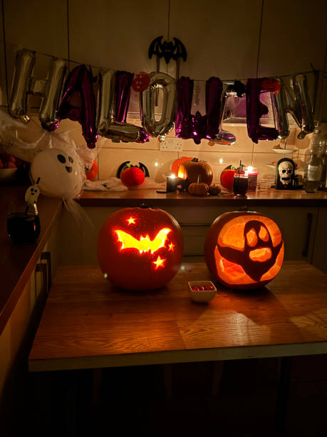 Stock photo showing a dimly lit kitchen decorated for Halloween themed party with silver and purple letter balloons spelling out the word 'Halloween' hanging from kitchen cupboard handles with illumination from candlelight. imagen de decoraciones de fiesta temáticas de halloween, cocina decorada para tallado de jack o' lantern, letras de globos plateados y morados que deletrean 'halloween', murciélagos negros, tallados, calabazas iluminadas en la mesa, luz de velas