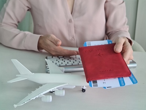 A womans hand holding a red passport with two plane tickets. On the white table is a toy airplane, symbolizing the concept of travel and tourism, adventure, exploration, and vacation planning.