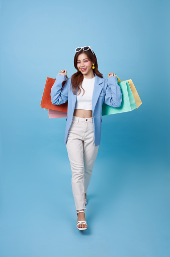 Full length young asian shopper woman walking and holding shopping bag feeling happy excited for discount promotion isolated on blue background.
