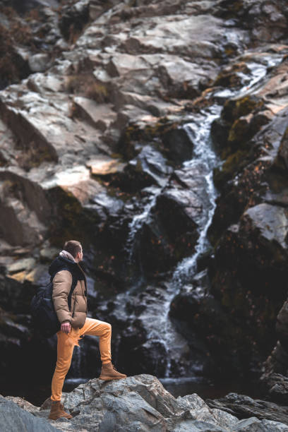 Young hiker man with backpack standing near to a waterfall and looking at it