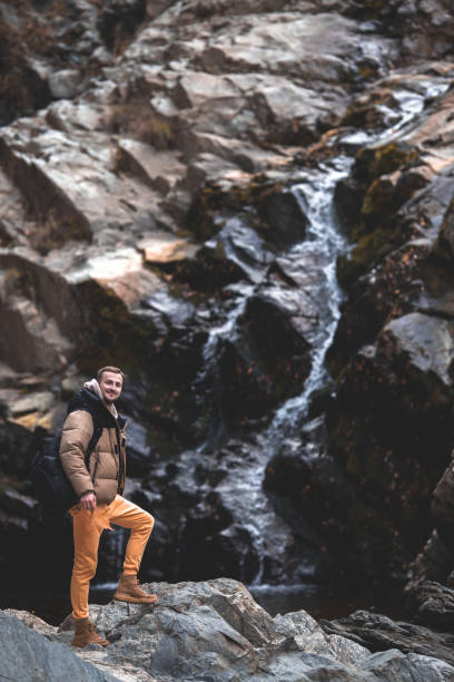 istock Young tourist man with backpack standing next to a waterfall and looking at the camera 1987299575