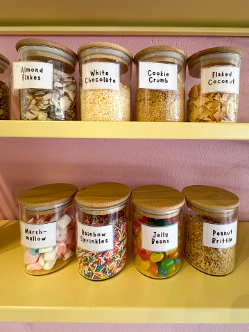 Stock photo showing close-up view of pantry shelves with an assortment of glass jars full of various baking ingredients for decorating cakes, including multicoloured, smarties sweet candy confectionary, marshmallows, peanut brittle, jelly beans, sugar sprinkles, cookie crumbs, flaked nuts such acs coconut, almonds and pistachios. These jars are lined up and organised neatly on shelves for ease of access, with labels on them.