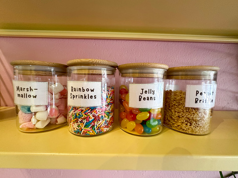 Stock photo showing close-up view of pantry shelves with an assortment of glass jars full of various baking ingredients for decorating cakes, including multicoloured, smarties sweet candy confectionary, marshmallows, peanut brittle and jelly beans. These jars are lined up and organised neatly on shelves for ease of access, with labels on them.