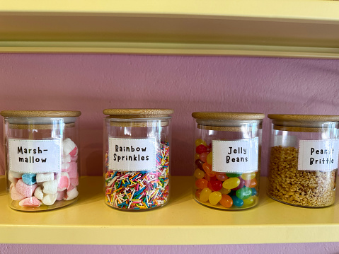 Stock photo showing close-up view of pantry shelves with an assortment of glass jars full of various baking ingredients for decorating cakes, including multicoloured, smarties sweet candy confectionary, marshmallows, peanut brittle and jelly beans. These jars are lined up and organised neatly on shelves for ease of access, with labels on them.