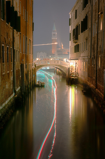 Night time in Venice and light trails from a small boat manoeuvre up a side canal towards the city's Grand Canal. In the distance we see the iconic shape of the Church of San Giorgio Maggiore.