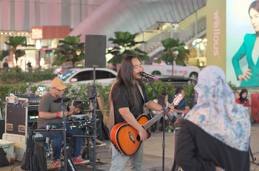 A man with long hair, singing and playing a guitar. Behind him is his bandmate on the drums. \n\n4 February 2024, Jalan Bukit Bintang, Kuala Lumpur, Malaysia