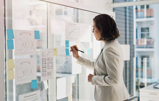 istock Business woman, writing and charts on board in office, planning and review for problem solving. Person, employee and strategy with notes for stats, analysis or reading for financial growth at company 1987369776