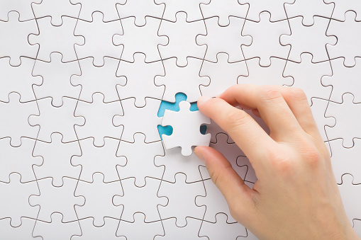 Young adult woman hand holding and putting last one white puzzle piece in middle on blue table background. Closeup. Top down view.