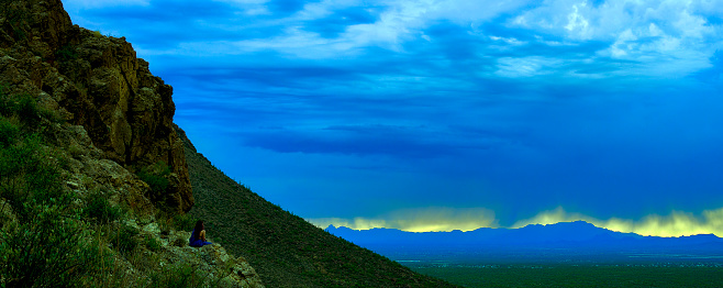 A person sits in solitude on a mountain edge at Gates Pass, Tucson, overlooking the vast landscape just after a summer storm at sunset.