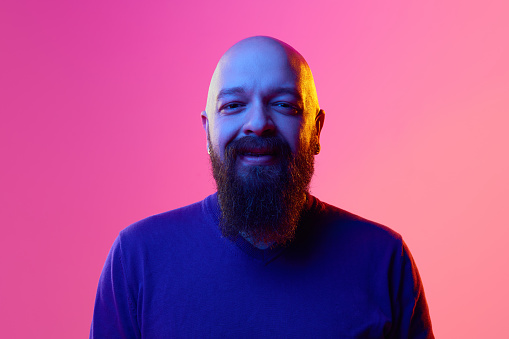Gel portrait of bearded bald man in his 30s wearing blue sweater, smiling, looking in camera against pink background in neon light. Concept of human emotions, facial expression. Positive mood