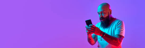 istock Bearded bald man in shit t-shirt emotionally looking on mobile phone against gradient purple background in neon light 1987397634