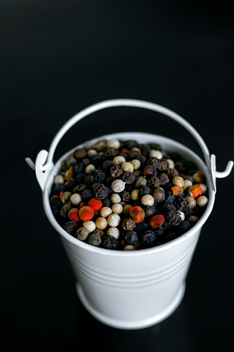 A mixture of pepper varieties with peas in the decorative little white bucket on the black background. Heap of various pepper. Mix of red, black, and white peppercorn seeds. Top view.