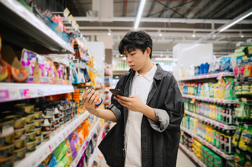 Asian Chinese Student Looking At Smartphone While Choosing Items In Supermarket.
