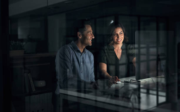 istock Night, man and woman at computer in agency for collaboration, software or creative project deadline. Graphic design, teamwork and happy business people coworking together in dark office for planning. 1987450343