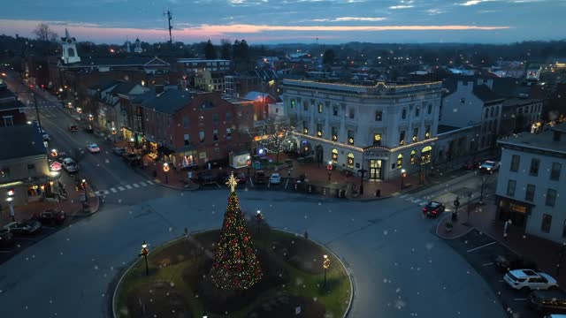 Snowflakes falling on small historic town in USA with Christmas tree and decorations at night. Aerial reveal.