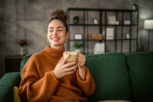 A young beautiful woman is sitting in a comfortable couch in her apartment and enjoying her morning coffee