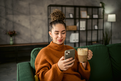 A young beautiful woman is sitting in a comfortable couch in her apartment and enjoying her morning coffee