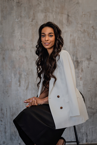 Smiling African American female portrait. Black long haired woman against gray wall. She is wearing black dress and white blazer. Copy space.