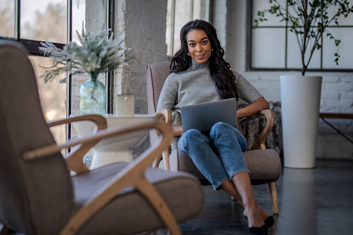 An attractive African American woman sitting in an armchair by the window and using her laptop. Smiling businesswoman working at the cafe. Full length shot.