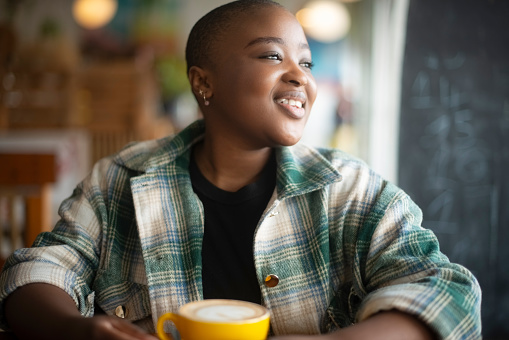 Black woman, idea and happy for coffee in restaurant, inspiration and tea break in morning for happiness. African person, smile and thinking by cup of cappuccino, wellness and warm beverage in cafe