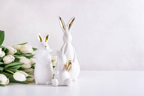 istock Three Easter rabbits figurines with bouquet of white tulips on white background. Easter celebration concept. Copy space. Front view 1987505276