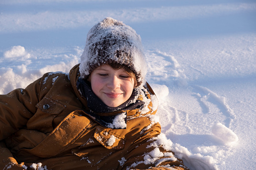 Portrait of smiling cute teenage boy, preteen playing in snow, in a snow-covered hat  in winter nature. Brown coat