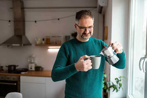 Contented mid-adult Caucasian man day standing near the window and holding moka pot while drinking coffee