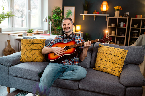 Portrait of an carefree mid-adult Caucasian man sitting on the sofa in his cozy home, and playing an acoustic guitar