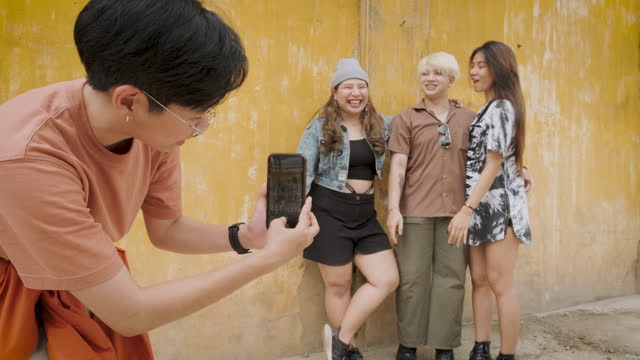 Group of young Asian people friends enjoy take a photo with mobile phone and standing at yellow old wall in background
