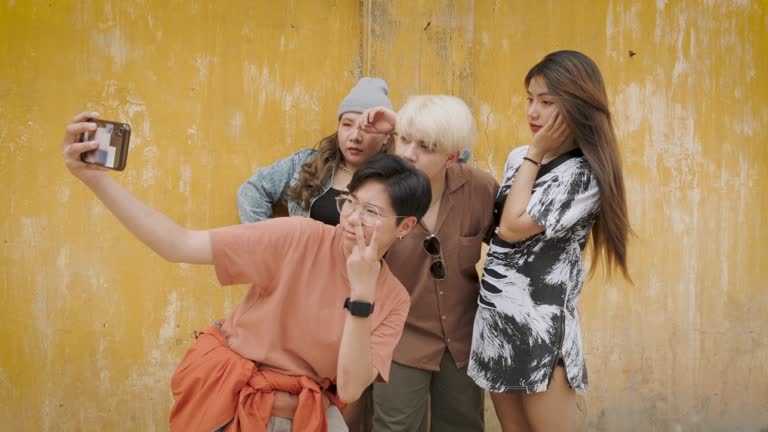 Group of young Asian people friends enjoy take a selfie with mobile phone and standing at yellow old wall in background