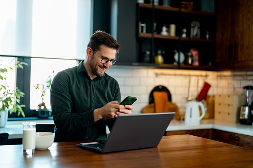 Smiling business man working at home with laptop and uses a smartphone