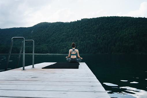 Woman meditating by the mountain lake on a summer day. Rear view of the woman sitting in a lotus position.