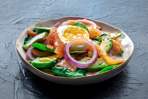 Salmon, avocado and egg salad with fresh leaves and onions, on a black stone background. Healthy diet