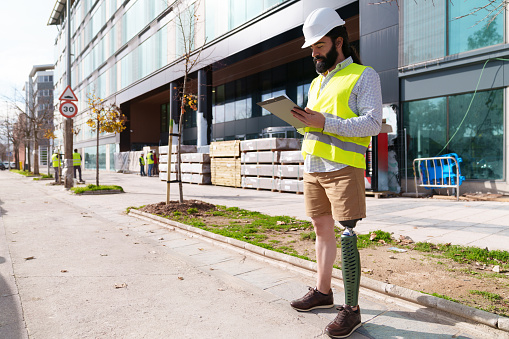 Construction worker with a prosthetic leg reviews digital blueprints on his tablet outdoors.