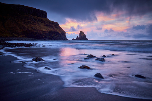 Talisker bay on Isle of Skye during a colorful sunset in December. There are rocks in the water and the sky is reflecting in the water.