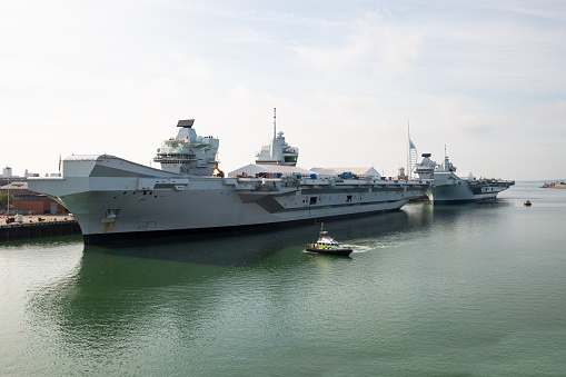 HMS Queen Elisabeth R08 (in front) and HMS Prince of Wales R09 docked at Portsmouth Naval Base.  Two police patrol boats survey the waters.