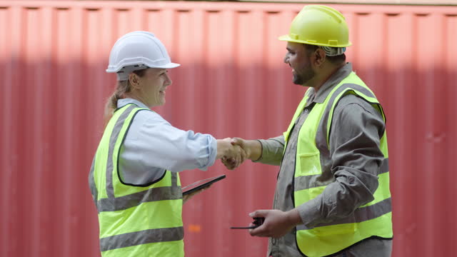 Happy dealing shaking hands two foreman worker working checking at container cargo harbor to loading containers, engineer man having hand shack with his woman boss after work discussion