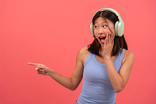 Beautiful young asian woman wearing headphones, pointing finger at the left side of the photo with a surprised face. The girl standing in the studio. Concept of advertising and presentation.