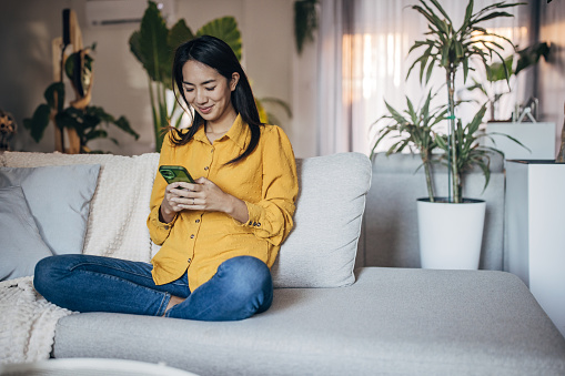 A cheerful young Japanese woman in a yellow shirt texts on her phone, sitting on the couch at home.