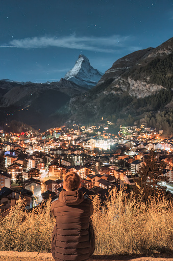 Rear view of man sitting and enjoying the view of illuminated Zermatt village and Matterhorn mountain in the night at Switzerland