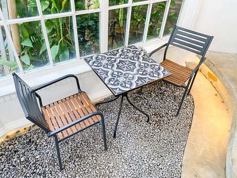 Cafe tables and chairs set made of black metallic frame and wood seat with gravel floor background near white framed glass window. Modern exterior or interior design concept for hotel, resort, villa.