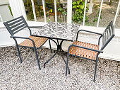 Cafe tables and chairs set made of black metallic frame and wood seat with gravel floor background near white framed glass window. Modern exterior or interior design concept for hotel, resort, villa.