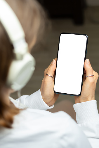 A woman in headphones holds a phone with a white screen listening to music. Smartphone mockup.