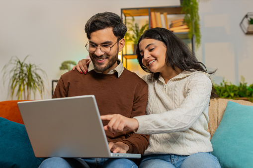Cheerful young diverse couple in casual clothes discussing while using laptop on sofa in living room at home. Happy Indian family planning together pointing showing booking online tickets in apartment