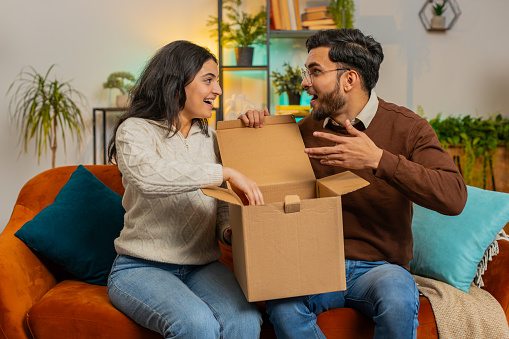 Happy excited couple open cardboard box together sitting on sofa at home. Hispanic young family consumers unpack good parcel looking inside giving high-five great purchase delivered by postal shipping