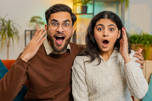 Portrait of amazed shocked couple with wow reaction open mouth looking at each other sitting on sofa in living room at home. Happy Indian family with big eyes, shocked by sudden victory, good win news