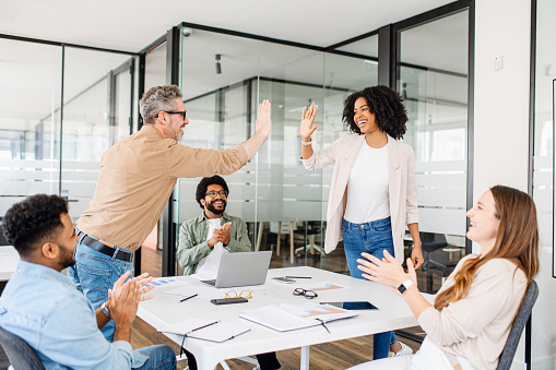 Elation and team spirit are palpable as two colleagues high-five in a contemporary office space, celebrating a collaborative achievement with vibrant enthusiasm. The diverse team applauds happily